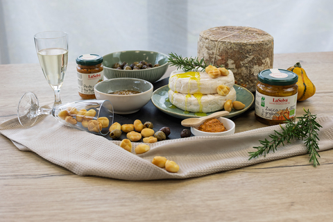 Das Bild zeigt eine ansprechend angerichtete Tapas-Platte mit verschiedenen Käsesorten, eingelegten Oliven, Pesto, einer Glasflasche, die einen Aperitif enthält, und Dekorationselementen wie einem Handtuch und Rosmarin.