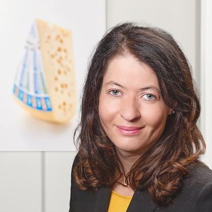 Das Bild zeigt eine Frau vor einem Hintergrund mit einem Stück Käse.