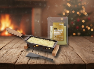 Das Bild zeigt ein Raclette-Gerät mit geschmolzenem Käse und einer Verpackung für Bio-Allgäuer Raclette, vor einem gemütlichen, festlichen Hintergrund.