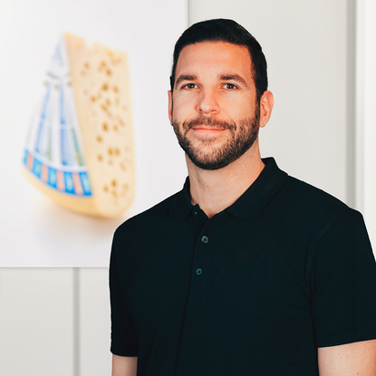 Auf dem Bild ist ein Mann in einem schwarzen Poloshirt vor einem Hintergrund mit einem Bild von Käse zu sehen.
