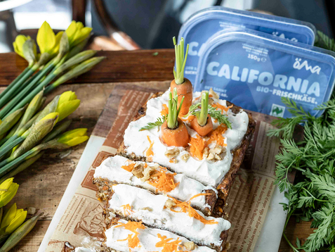 Das Bild zeigt einen bunten Karottenkuchen, der mit Frischkäse, geraspelten Karotten und kleinen Karotten-Dekorationen garniert ist, umgeben von frischen Kräutern und Blumen.
