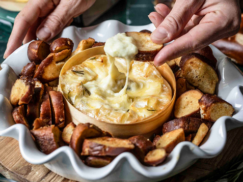 Auf dem Bild ist ein käsiger Dip zu sehen, der mit knusprigem Brot serviert wird, während jemand ein Stück Brot in den Dip taucht.